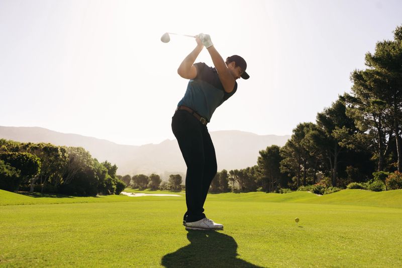 A man golfer skillfully swings his club during a fairway shot, with a golf ball in sight, surrounded by vibrant greenery under a clear sunny sky.