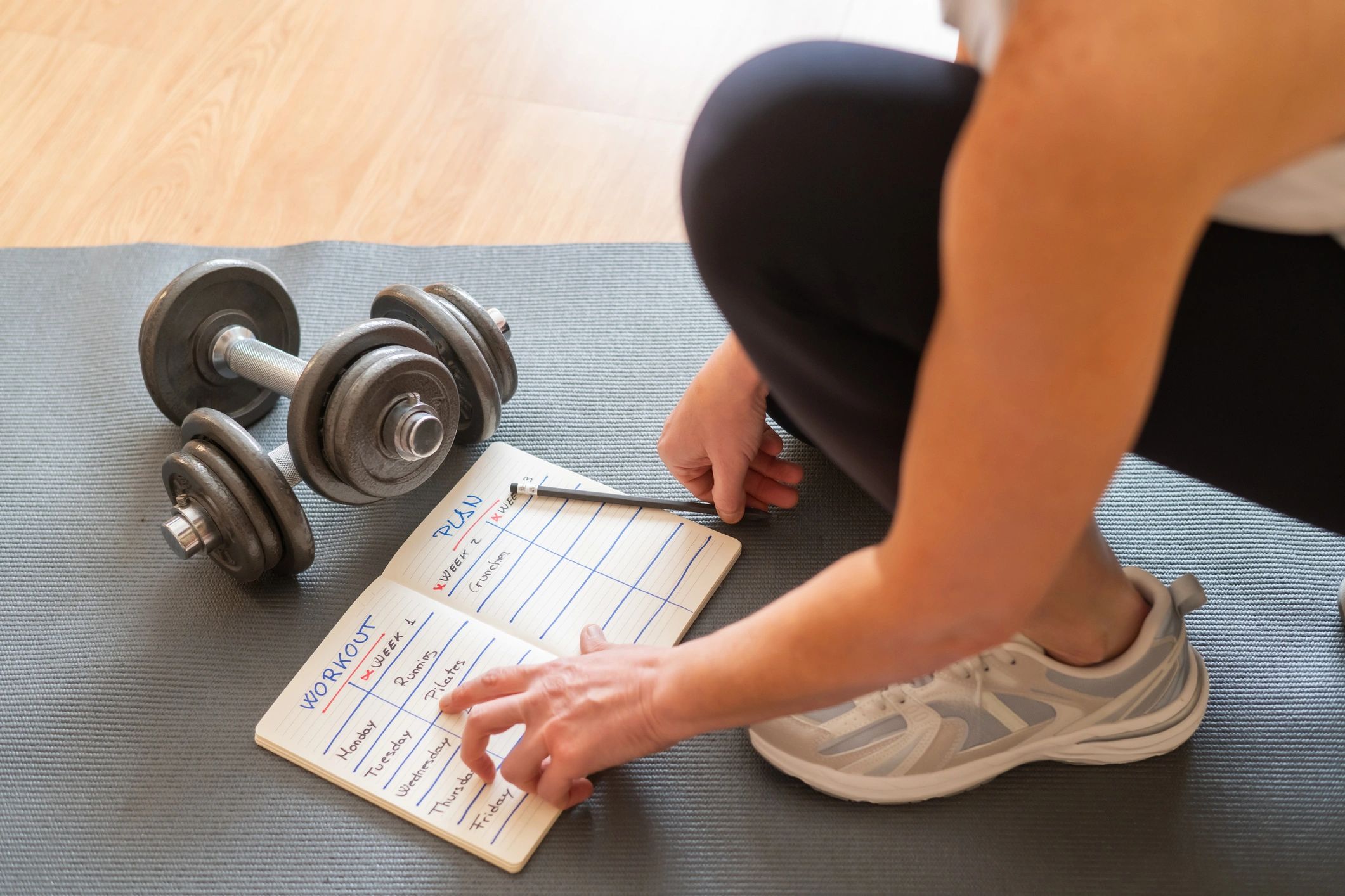 Person checking workout plan next to dumbbells on mat.