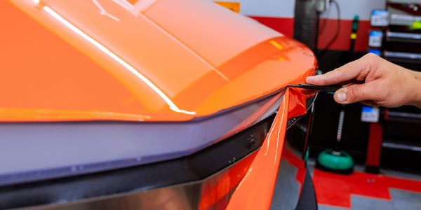 A hand applying orange vinyl wrap to a car's rear end.