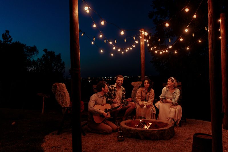 Friends enjoying guitar music under string lights, smiling and having fun next to a campfire on a summer bight.