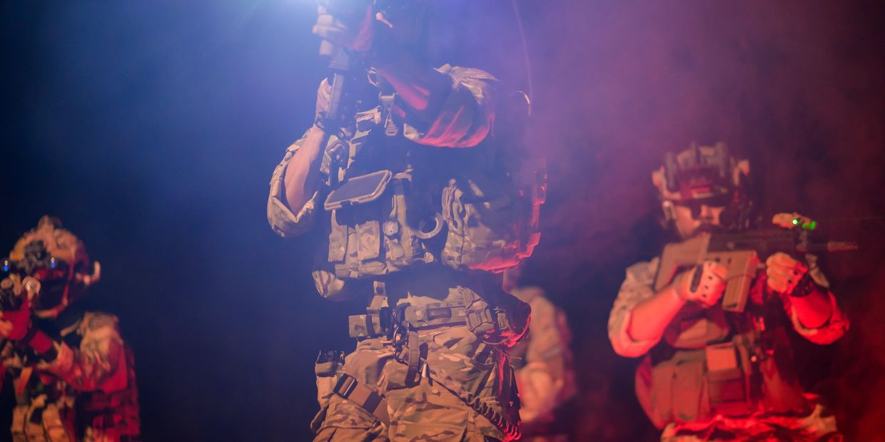 Soldiers in tactical gear aiming weapons at night with red and blue lighting.