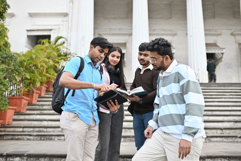 A group of university student friends standing together on the steps of the campus, engaged in studying and discussing their notes. With books in hand, they share ideas and collaborate in a relaxed, outdoor setting. The vibrant campus surroundings reflect a moment of academic focus, teamwork, and friendship among students.
