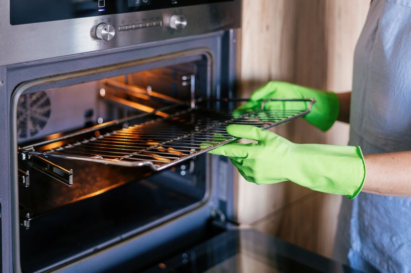 A woman wearing green cleaning gloves carefully remove a dirty rack from a modern oven. The kitchen features and household appliances concepts. Electric equipment for home