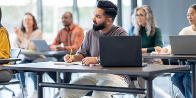A student attentively taking notes in a classroom with laptops and classmates around.