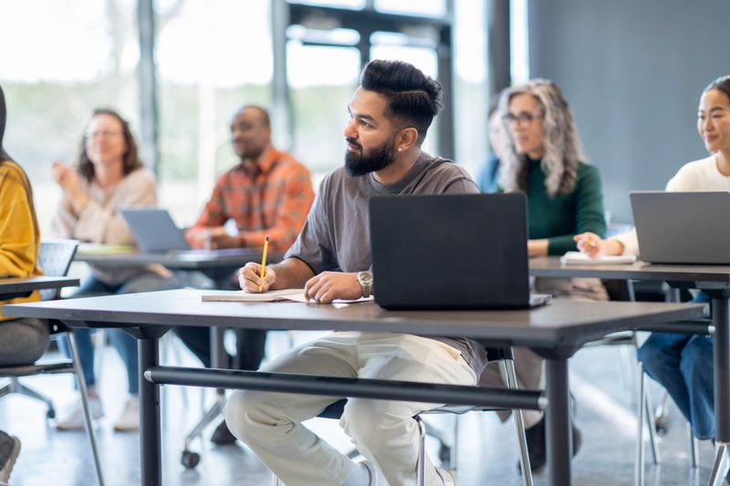 A large group of mature adults are seen sitting at desk with wither laptops and books open as they study during a continuing education class.  They are each dressed casually and are listening attentively.
