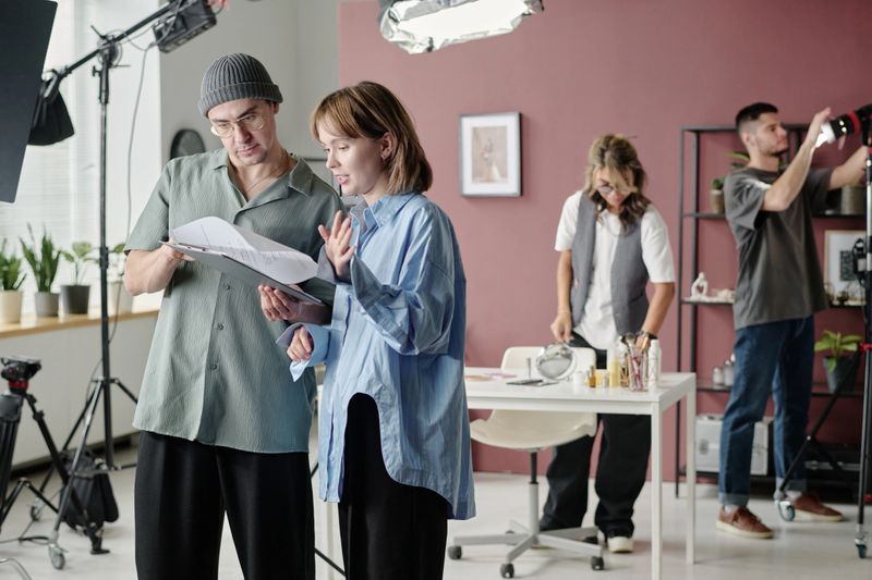 Caucasian director and actress reading script together while production designer and operator checking props and light in blurred background