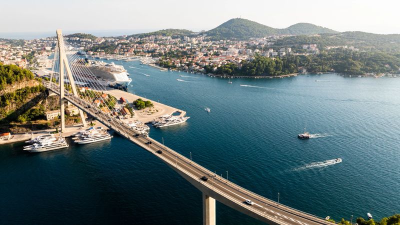 The Franjo Tu?man Bridge Stretches Over the Sparkling Sea,Framed by Towering Mountains and a Charming Coastal Town. Ships Glide Beneath the Bridge,Adding Life and Movement to the Picturesque Scenery
