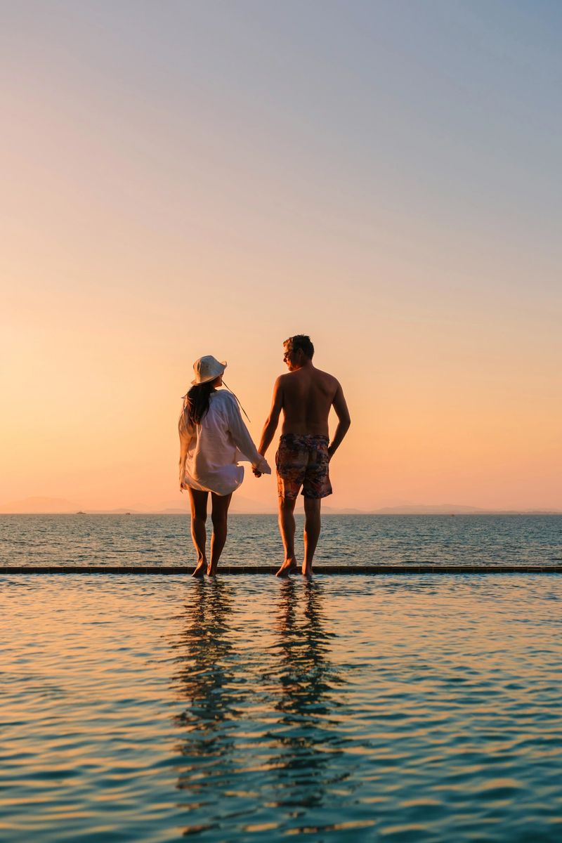 Couple of men and woman enjoy a romantic moment, holding hands while walking along the edge of a serene pool, with the golden sunset reflecting on the tranquil waters of Koh Munnork Island in Thailand