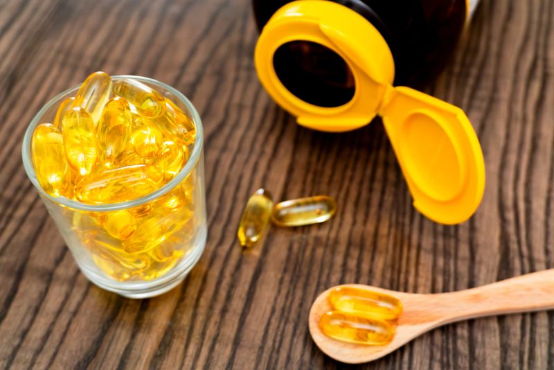 A glass filled with yellow gel capsules sits beside an open bottle on a wooden surface. A wooden spoon leans next to the glass, highlighting the contents.