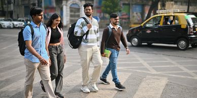 Four young people walking on a crosswalk in an urban area with a yellow-black taxi in the background.