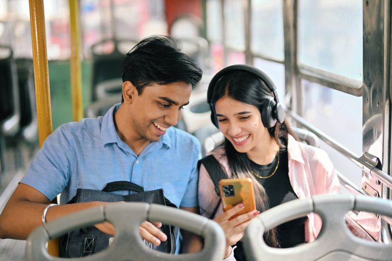 A young man and woman, university students, commuting on a bus, seated together and smiling as they look at a mobile phone. They are casually dressed, enjoying a quiet moment, reflecting connection and modern lifestyles in an urban setting