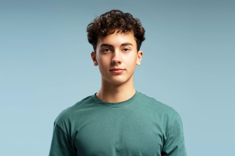 Portrait of handsome teen boy with curly hairstyle wearing green t shirt looking at camera, posing isolated on blue background. Advertisement concept