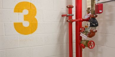 Red fire sprinkler pipes and valves beside a large yellow number 3 on a white brick wall.