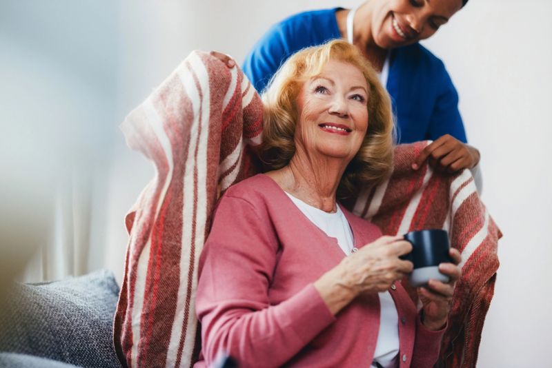 An elderly woman relaxes with a caring caregiver wrapping a blanket around her shoulders, portraying comfort and support. The scene reflects warmth, compassion, and the connection between caregiver and recipient.
