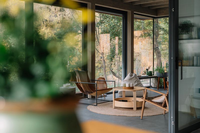 Photo of modern wooden cabin interior featuring a minimalist dining area bathed in natural light. Large glass windows provide a serene view of the forest with vibrant autumn foliage, creating a peaceful nature-inspired atmosphere. The clean design includes wooden furniture, a spacious table, and contemporary chairs, offering a tranquil and cozy retreat perfect for relaxation or a getaway. Ideal for concepts of modern living, eco-friendly homes, sustainable architecture, and indoor-outdoor harmony