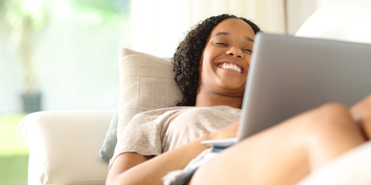 A smiling woman relaxing on a couch with a laptop.