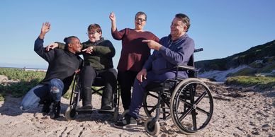 Group of friends, including two in wheelchairs, enjoying a sunny beach day together.