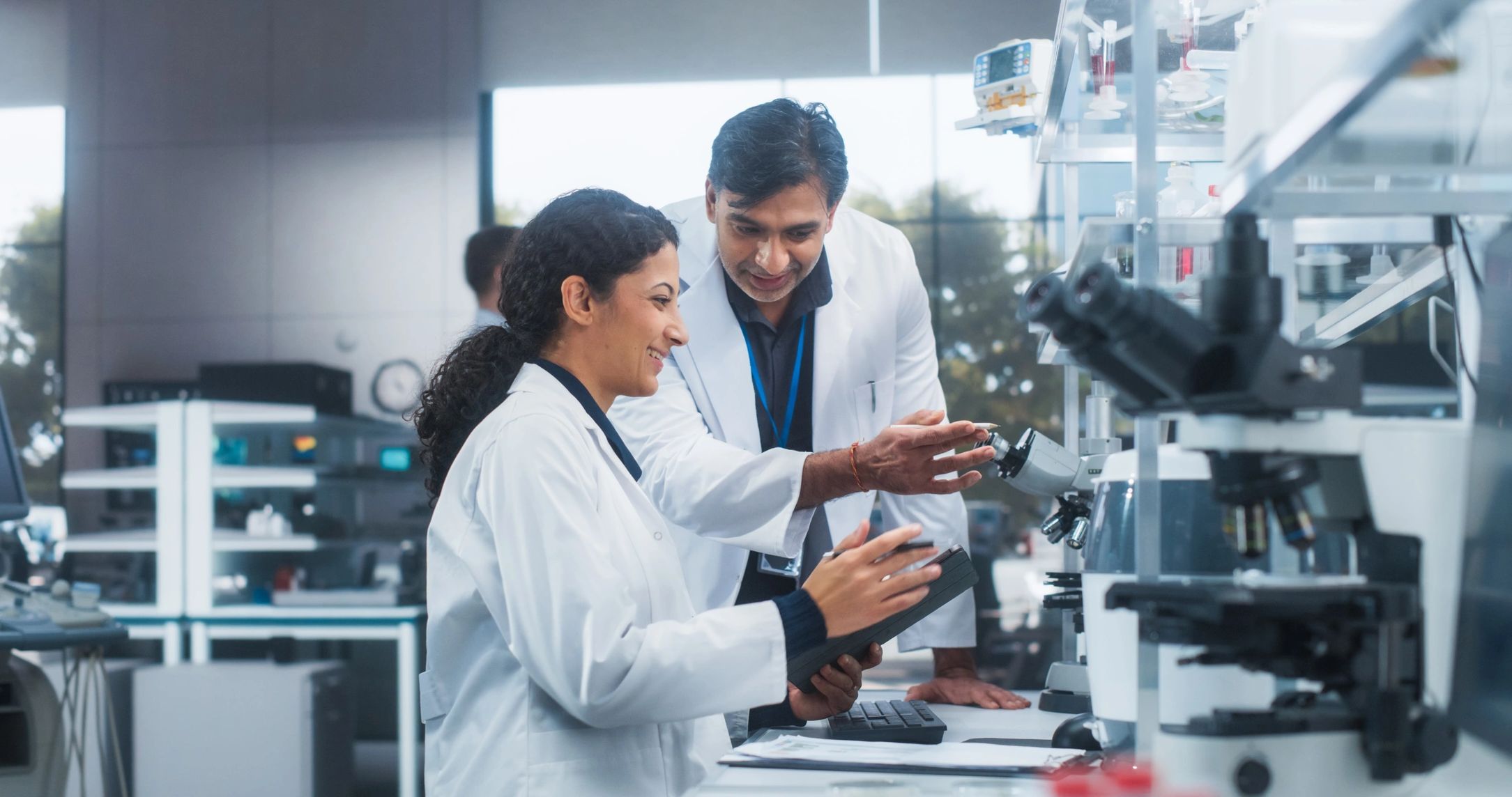 Two scientists in white lab coats working together with a microscope in a modern lab.