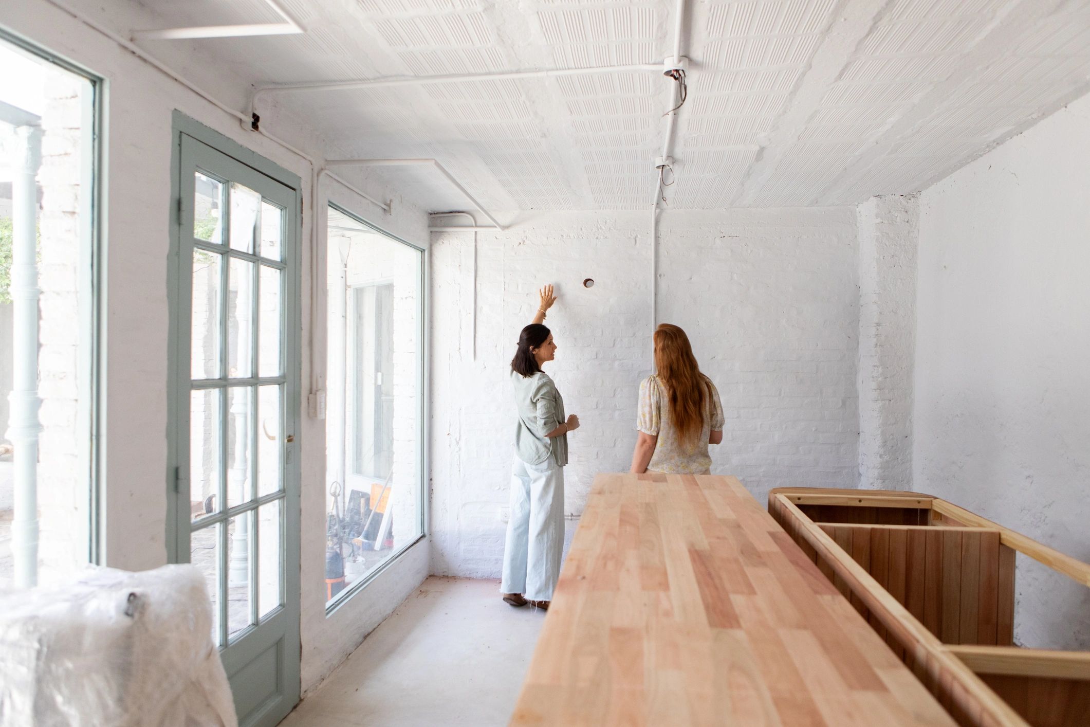 Two women discussing renovation plans in a bright, empty room with wooden countertops.