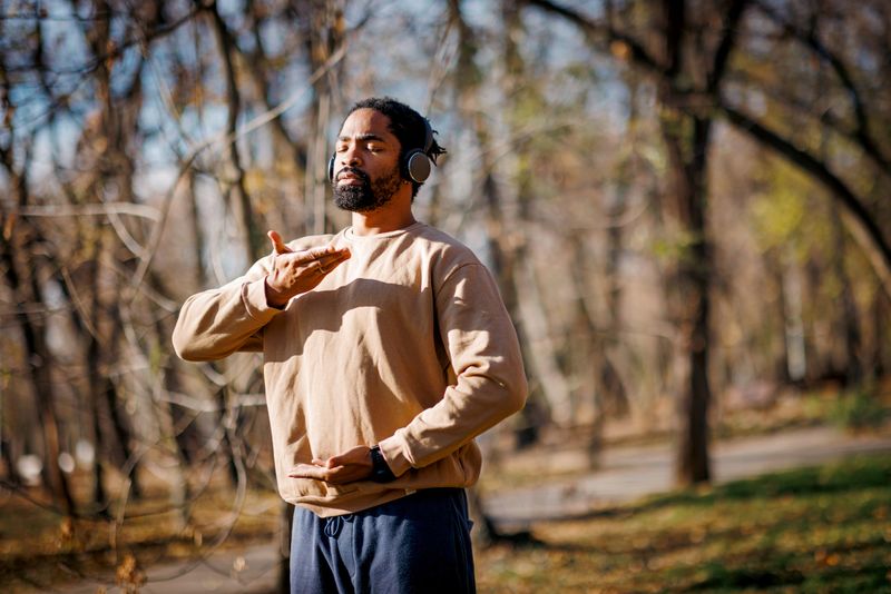 Young adult man practicing qigong in a public park, wearing headphones and soaking in the warm sunlight filtering through vibrant autumn trees, embracing tranquility and relaxation