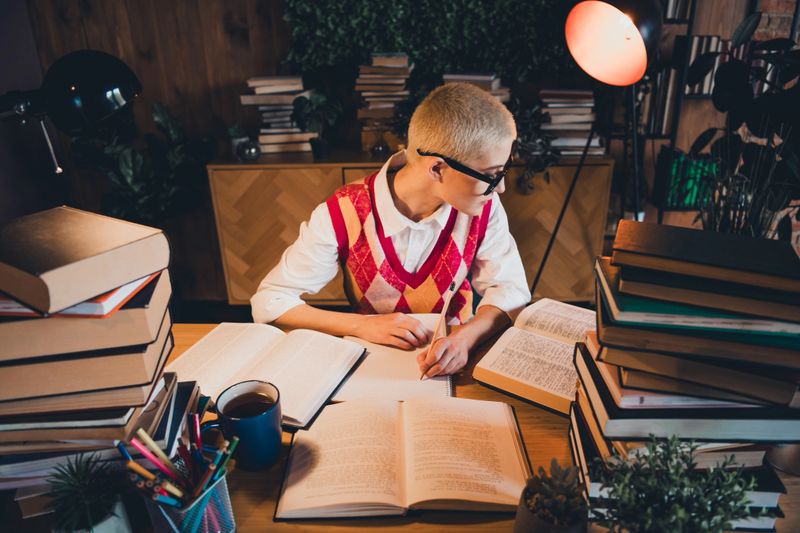 A young woman with short blond hair and stylish glasses studies late in the evening. Her argyle waistcoat complements her casual style, creating a cozy atmosphere with books and natural lighting.