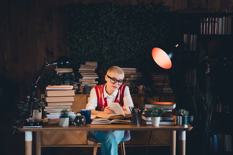 A young woman with short blond hair and glasses sits at a wooden desk, studying in a softly lit home library, surrounded by books and plants.