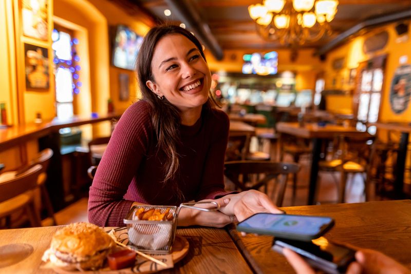 Cheerful young woman is paying for her meal using contactless payment with her smartphone in a cozy restaurant, enjoying a delicious burger and fries