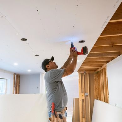 A worker installs drywall on a ceiling using a power drill.
