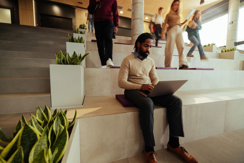 Focused businessman using laptop in modern office lobby, ignoring busy colleagues passing by