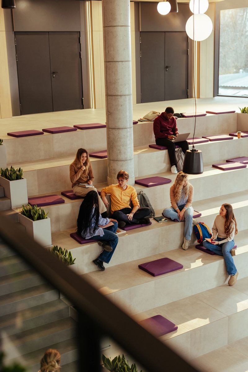 Diverse group of university students are sitting on steps in a modern open space area, studying and socializing between classes