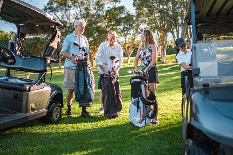Two mature diverse couples prepare for golf in stylish attire on a sunny course. The men handle golf bags while the women select clubs. Golf carts and lush trees complete the serene golfing environment.