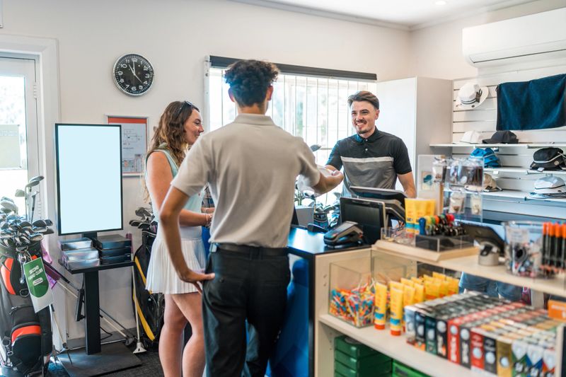 Young adult Caucasian male golf store seller enthusiastically welcomes diverse customers in a well-stocked retail setting. Dressed in a professional attire, he assists with purchases amid shelves of golfing equipment and merchandise in a modern golf shop.