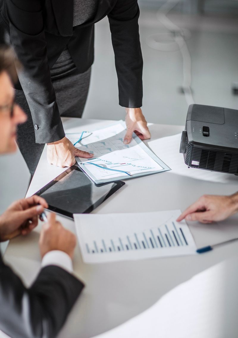 A diverse group of professionals examines financial reports in a modern office. The team, dressed in business attire, utilizes advanced technology, including tablets and projectors, to enhance their strategic planning during the meeting.