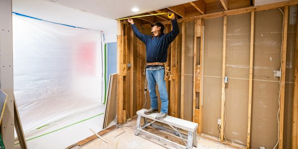 Man measuring space in wood-framed room under construction.