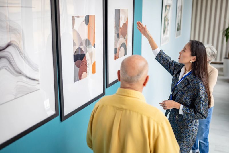 An art gallery curator guides visitors through a collection of framed modern artwork, explaining the pieces on display. The setting portrays engagement with visual art in a contemporary and creative environment.