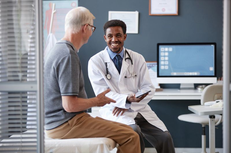 Waist up portrait of smiling African American doctor wearing lab coat and holding tablet while talking to senior patient during consultation in clinic, copy space