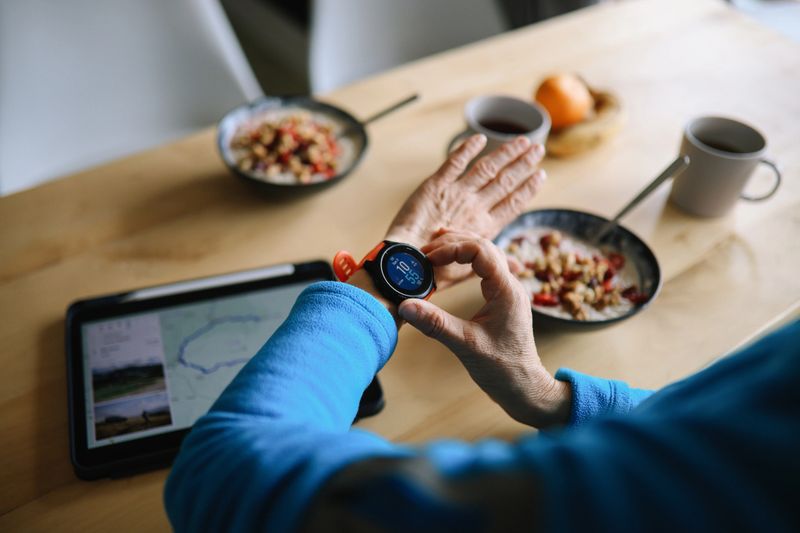 Woman in her 50s having a light breakfast while exploring running trails on her app and setting up a smartwatch for the exercise in the outdoors.