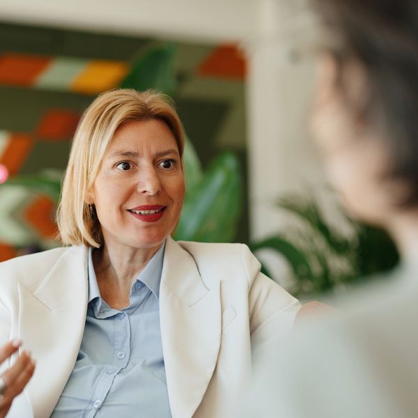 A blonde woman in a cream jacket is talking to another woman face ot face in an office.