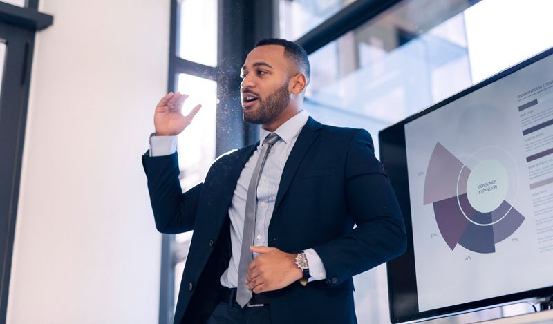 A confident business professional passionately delivers a presentation in a modern office setting. Dressed in a suit, he gestures energetically beside a screen displaying graphs. The image captures the essence of leadership, communication, and professional engagement, evoking a sense of ambition and focus.