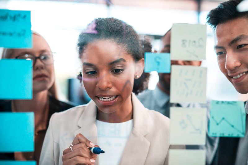 A diverse group of professionals brainstorming and planning together using sticky notes on a glass wall. This image conveys teamwork, innovative thinking, collaboration, and productivity in a modern office environment.