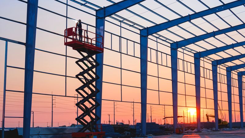 Silhouette factory building framework with welder worker on electric scaffolding working overtime to welding metal wall structure in construction site area against sunset sky background
