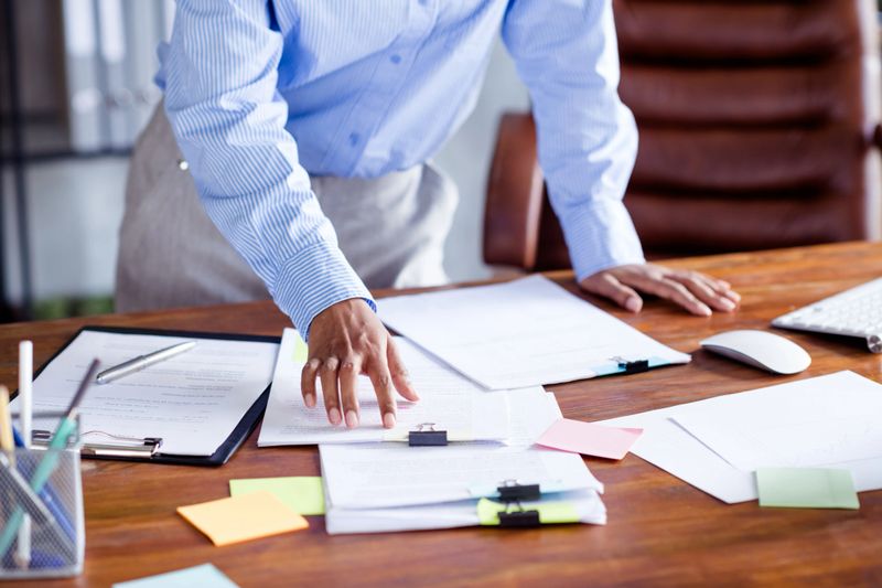 A young professional woman in a formal blouse reviews documents at her office desk, surrounded by paperwork, notes, and a computer. The setting conveys a sense of business focus and organization.