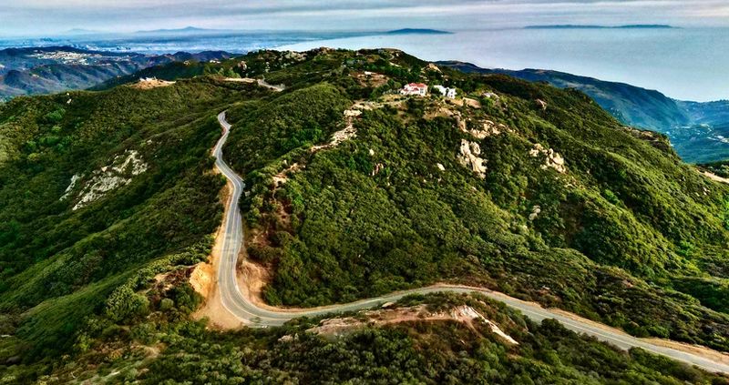 A sweeping panoramic aerial view of Santa Monica and Los Angeles from atop Santa Monica Mountains in Malibu with a windy road in foreground.