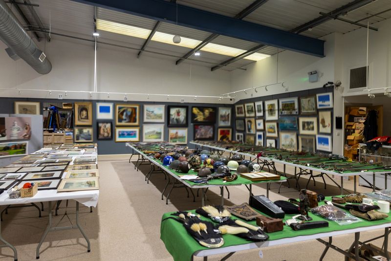 Wide shot of a large room in an auction house filled with various antiques and paintings. There are tables with war antiques such as helmets and swords.