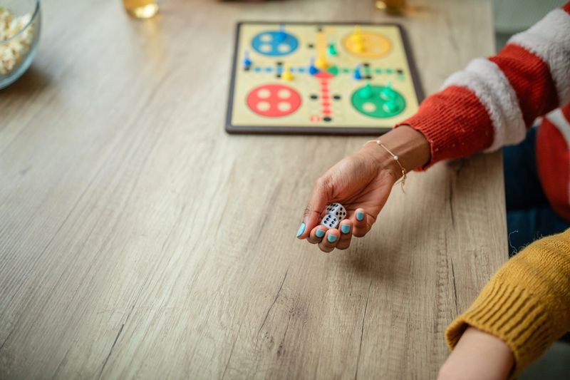 A person in a red and white sweater holds dice beside a colorful board game on a wooden table, ready to roll in a friendly, competitive setting.