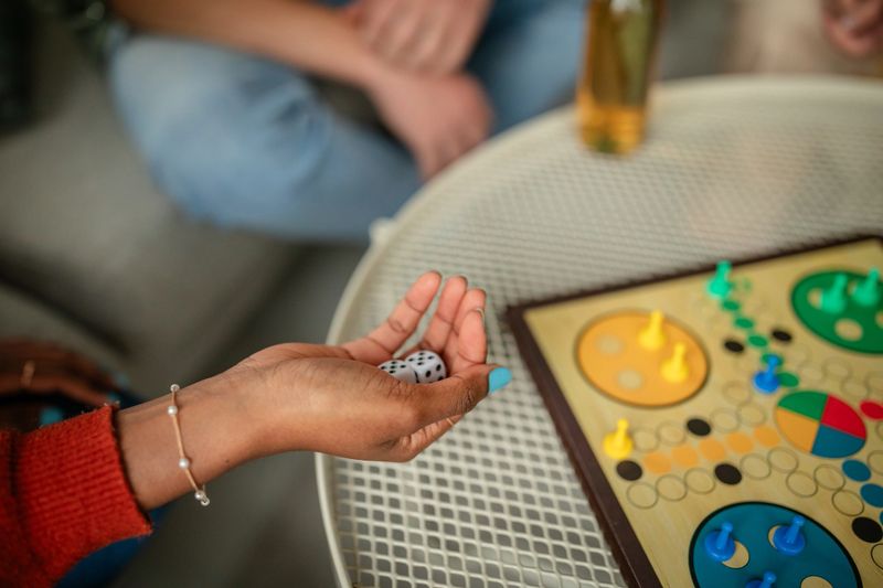 A close-up shows a hand holding dice, poised to make a move in a board game. Enthusiasm fills the air as drinks sit nearby, creating a lively and interactive atmosphere.