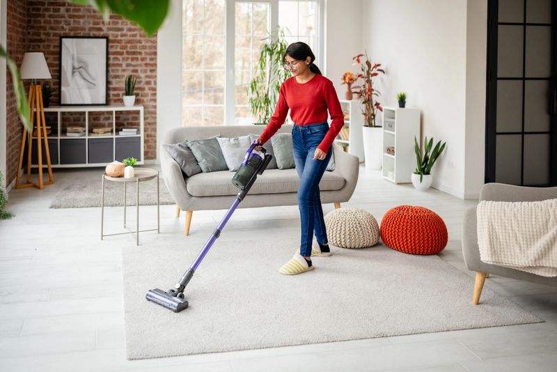 A woman in a red sweater and blue jeans is vacuuming a spacious, light-filled living room adorned with modern decor and soft furnishings, including colorful cushions.