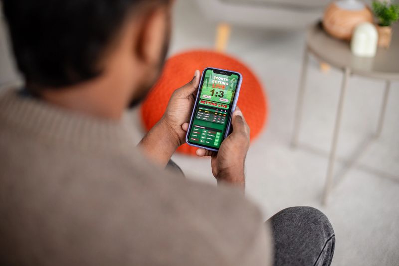 A man is engrossed in viewing the latest sports results or a mobile game on his phone screen, showcasing a modern lifestyle in a cozy home setting.