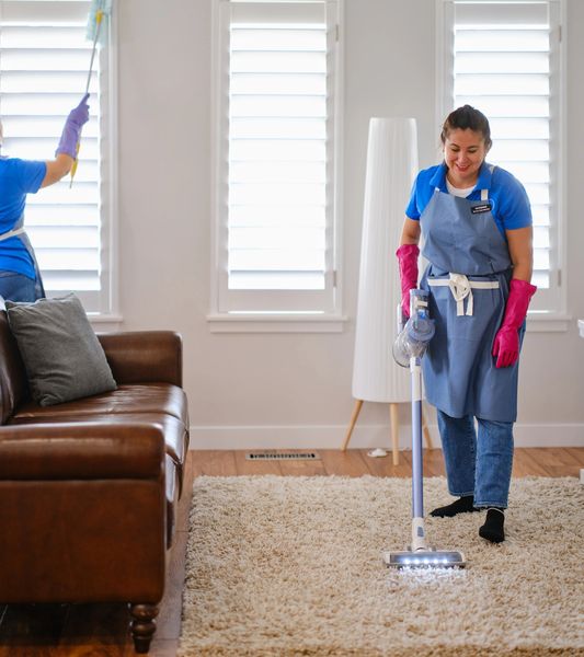 Two women cleaning a living room with vacuum and duster.