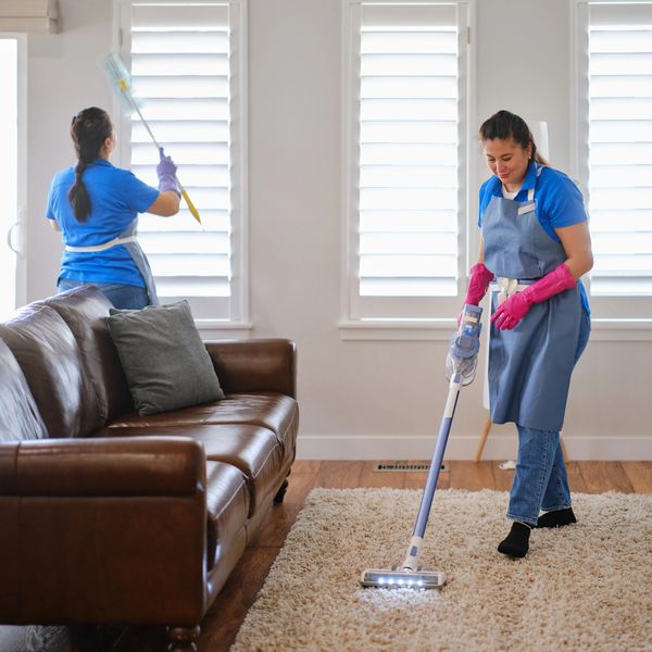 Two women cleaning a living room with vacuum and duster.